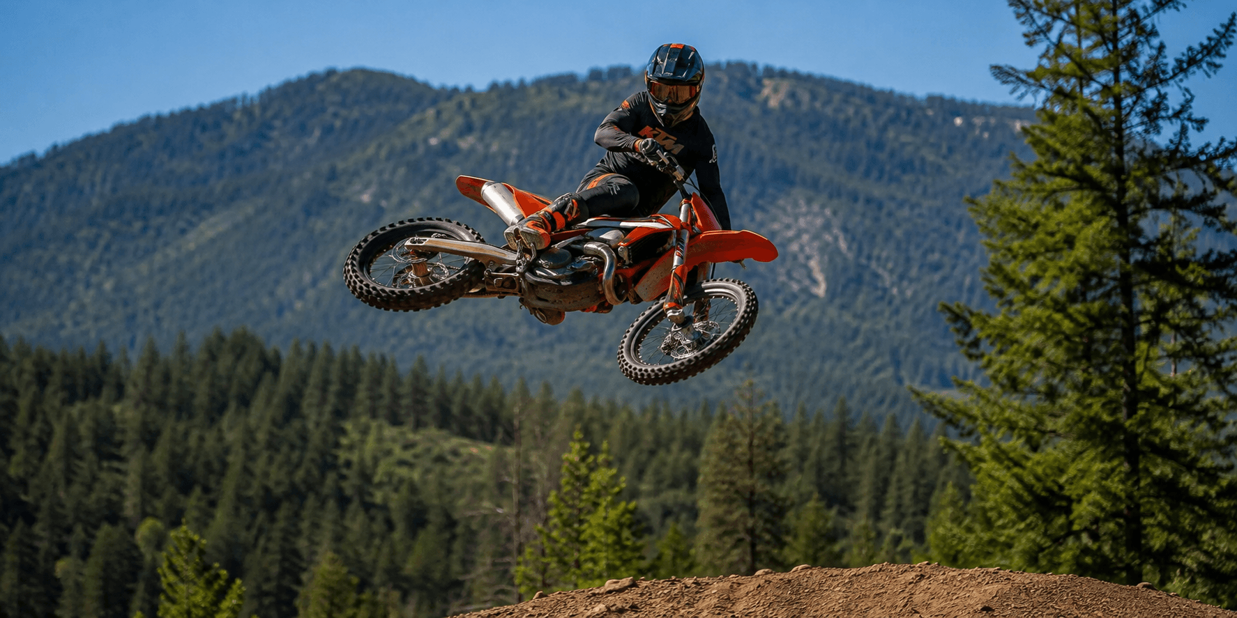 Dirt bike rider jumping a motocross track in front of a forested mountain